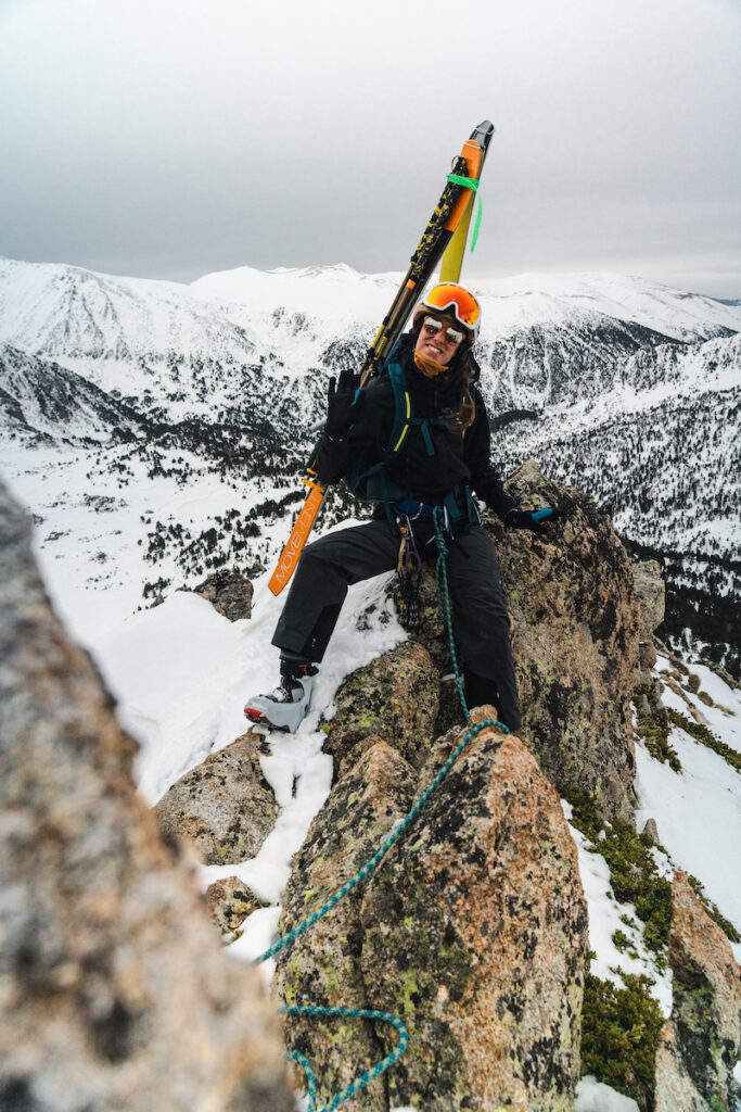 A woman with skis smiling on top of a snowy rocky mountain