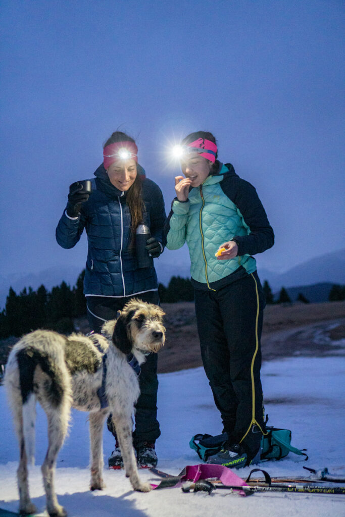Two female athletes fueling themselves in the snow covered outdoors with a dog in front of them looking at the camera