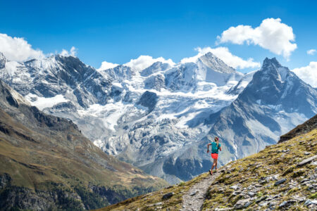 Female athlete trail running the Via Valais wearing bright orange shorts and a teal backpack and shirt.