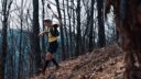 Trail runner John Kelly wearing a yellow shirt and black shorts running downhill through a forest full of fallen leaves and branches.