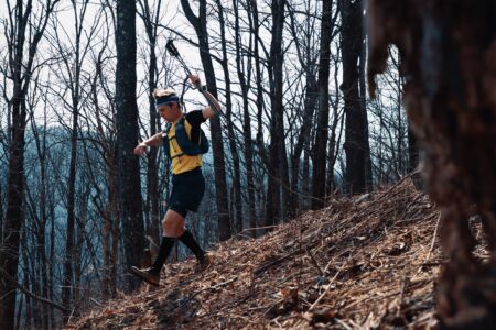 Trail runner John Kelly wearing a yellow shirt and black shorts running downhill through a forest full of fallen leaves and branches.