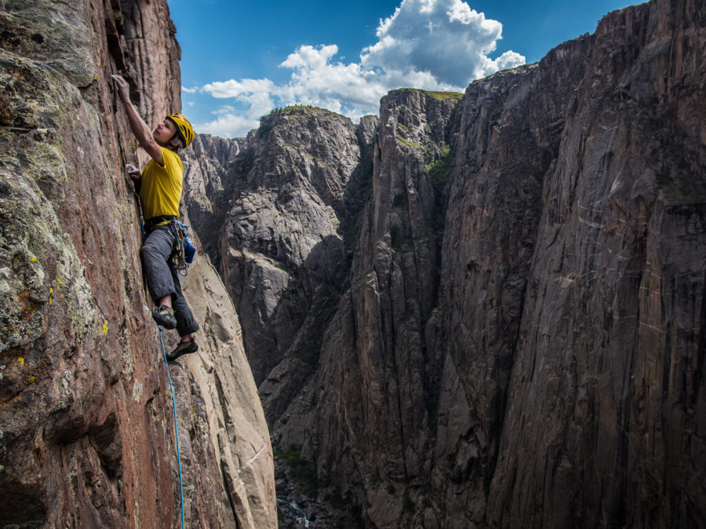 A climber in a yellow t-shirt leaning on a wall with his left hand looking for a hold.