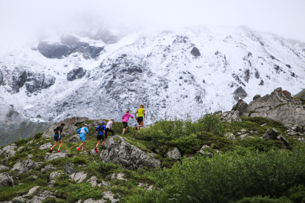 Six runners ascending uphill on a rocky terrain with a massive snow mountain in the background.