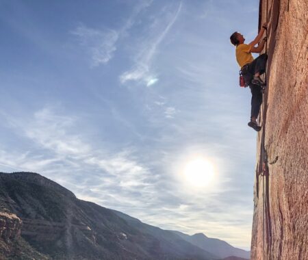 A man in a yellow t-shirt and blue pants climbs a straight wall with a chalk bag and rock climbing shoes.