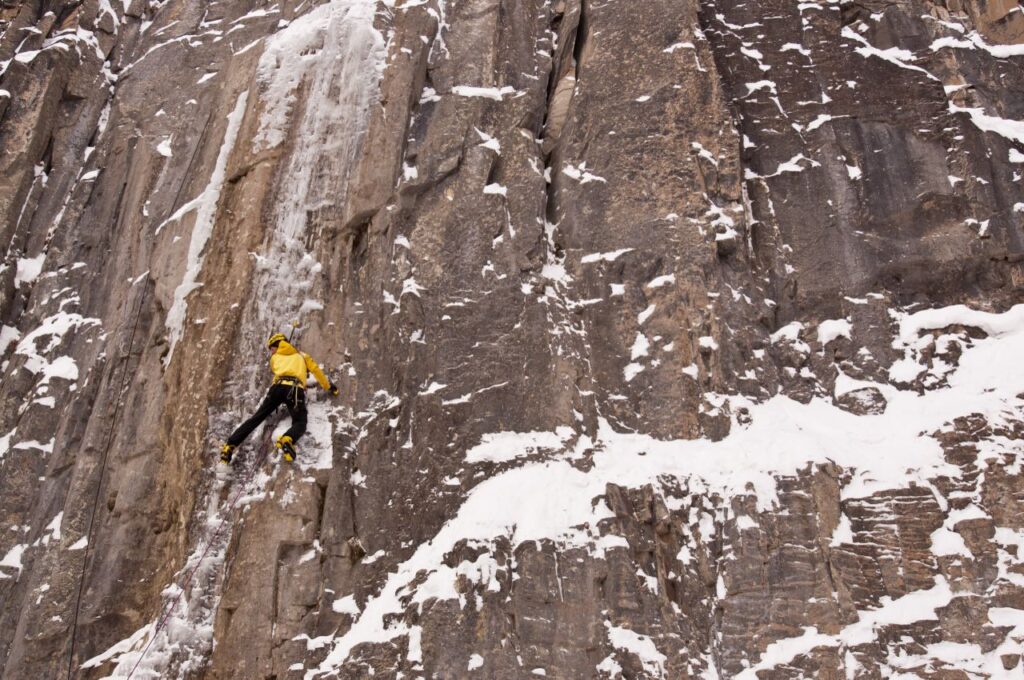 A climber in a yellow jacket and black trousers climbs a wall in snow and icy conditions.