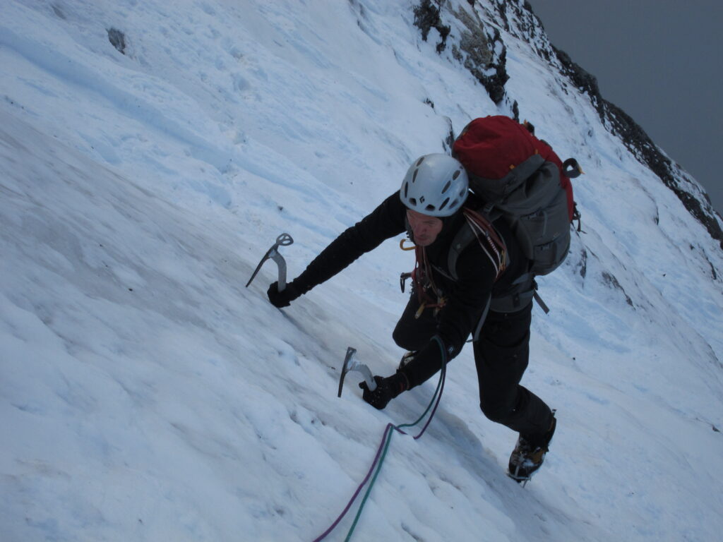 A climber scaling a snow-covered slope of a steep mountain with ice axes.