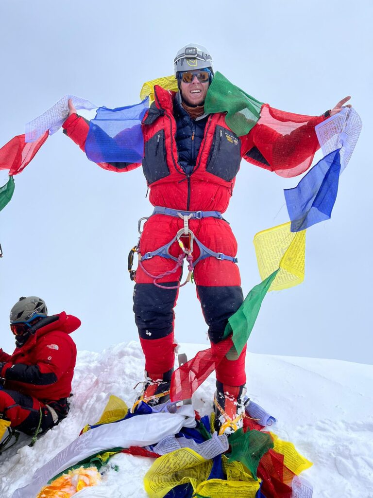 A male climber atop a mountain summit with Tibetan prayer flags around.