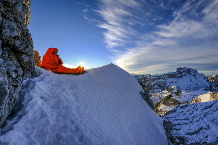 A man in a sleeping bag on an snowy mountain at sunset.