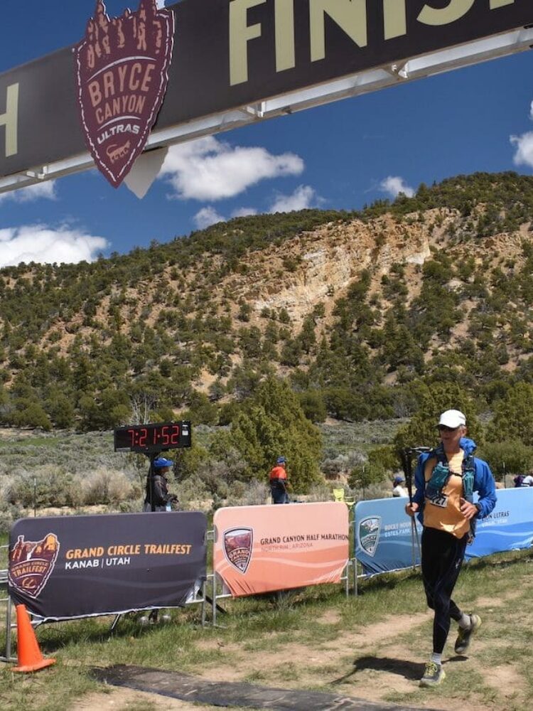 A male runner wearing a white hat crossing the finish line of a 100-mile race with poles in his right hand.